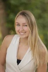 Eine junge Frau mit langen blonden Haaren und blauen Augen lächelt und schaut direkt in die Kamera. Sie trägt ein weißes ärmelloses Oberteil.
