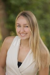 Eine junge Frau mit langen blonden Haaren und blauen Augen lächelt und schaut direkt in die Kamera. Sie trägt ein weißes ärmelloses Oberteil.