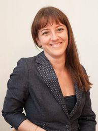 Eine Frau mit braunen Haaren und Ponyfrisur lächelt und trägt einen schwarzen Blazer mit weißen Punkten.