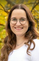 Eine Frau mit braunen, welligen Haaren und einer Brille lächelt in die Kamera. Sie trägt ein weißes Shirt und eine Halskette. Der Hintergrund ist verschwommen mit gelben Blättern.