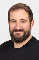 Ein Mann mit Bart und Schnurrbart lächelt in die Kamera, trägt ein schwarzes T-Shirt. Er hat kurze, dunkle Haare und einen sauber rasierten Kopf.