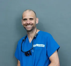 Ein Mann in einem blauen OP-Kleid mit einem Stethoskop um den Hals lächelt für ein Foto.