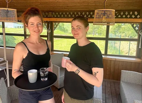 Zwei Frauen stehen in einem gemütlichen Raum, eine hält einen Tablett mit einer Tasse und einem Glas Wasser, die andere hält eine rosa Tasse.