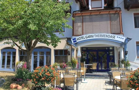 Die Vorderseite des Hotel Garni Thermenoase, ein Hotel mit blauen Fenstern und Türen, ist von Sitzgelegenheiten im Freien mit Tischen und Stühlen umgeben. Der Eingang ist durch ein blaues Vordach mit weißem Text und einem geschlossenen weißen Schirm gekennzeichnet.