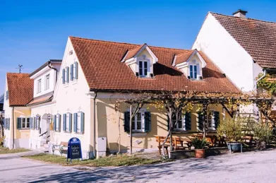 Ein gelbes Haus mit einem braunen Dach und blauen Fensterläden. Das Haus hat eine Holzbank und einen Tisch draußen.