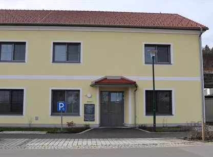 Ein gelbes Gebäude mit rotem Dach und weißer Umrandung hat ein Behindertenparkplatzschild, eine graue Tür und Fenster mit Jalousien.