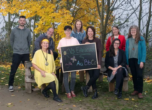 Eine Gruppe von Erwachsenen in einem Park lächelt und posiert für ein Foto. Zwei Frauen halten ein Kreidetafel mit den Texten 'VS Edelsbach' und '344 Aa BbCc'.