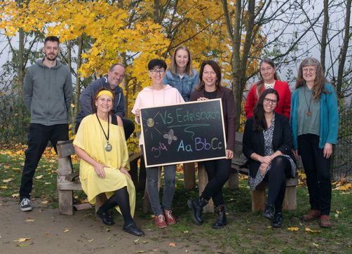Eine Gruppe von Erwachsenen in einem Park lächelt und posiert für ein Foto. Zwei Frauen halten ein Kreidetafel mit den Texten 'VS Edelsbach' und '344 Aa BbCc'.