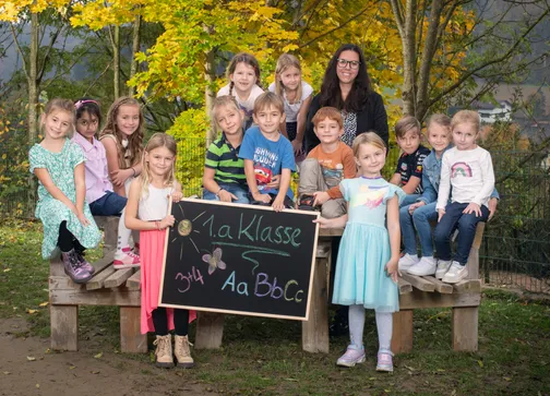 Eine Gruppe von Kindern, einige halten ein Tafel, posieren für ein Foto im Freien. Die Tafel steht '1.a Klasse'. Sie sitzen auf einer Bank, mit einer Frau vor ihnen. Der Hintergrund zeigt Bäume mit gelben Blättern.
