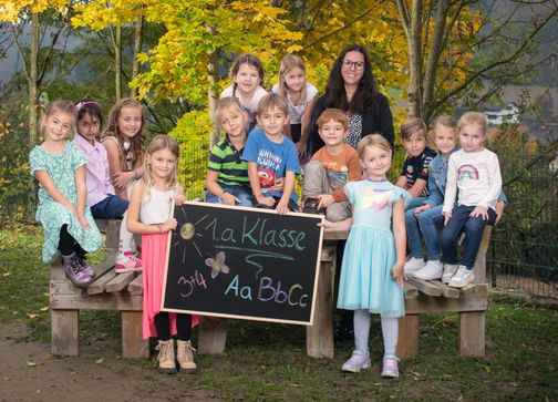 Eine Gruppe von Kindern, einige halten ein Tafel, posieren für ein Foto im Freien. Die Tafel steht '1.a Klasse'. Sie sitzen auf einer Bank, mit einer Frau vor ihnen. Der Hintergrund zeigt Bäume mit gelben Blättern.