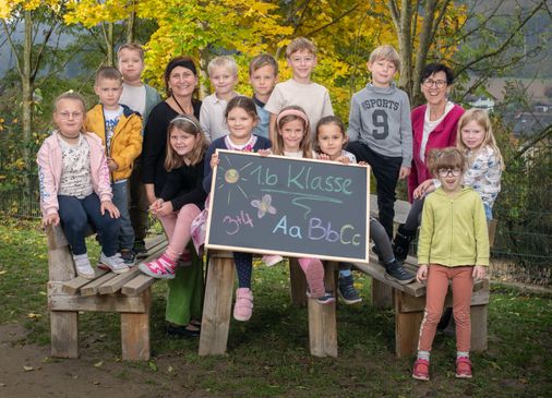 Eine Gruppe von Kindern, Lehrern und Eltern posiert für ein Foto im Freien. Die Kinder halten ein Tafel mit den Worten '1.b Klasse' und einer gezeichneten Schmetterling. Sie sitzen auf Bänken mit einem Grasbereich im Hintergrund.