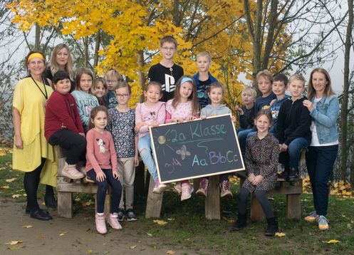 Eine Gruppe von Kindern und einer erwachsenen Frau posiert für ein Foto in einer Außenumgebung mit gelben Bäumen. Die Kinder halten ein Tafel mit dem Text '2a Klasse' und Namen.