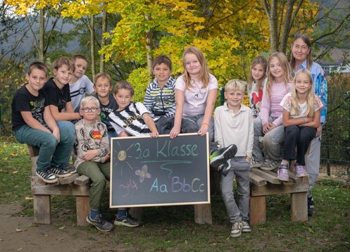 Eine Gruppe von Kindern und Erwachsenen sitzt auf einer Bank im Park und hält eine Tafel mit der Aufschrift '3a Klasse'. Die Kinder lächeln und scheinen für ein Foto zu posieren.