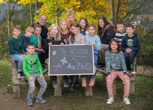 Eine Gruppe von Kindern und zwei Erwachsenen posiert für ein Foto. Die Kinder halten ein Tafel mit einer Nachricht. Im Hintergrund befinden sich Herbstbäume.