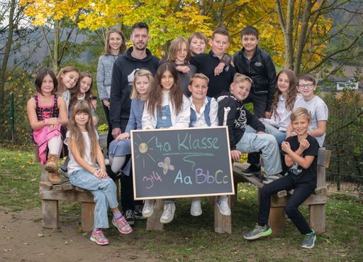 Eine Gruppe von Kindern und Erwachsenen posiert für ein Bild im Freien. Sie sitzen auf Holzbänken. Ein Tafel mit '4a Klasse' und '314' wird von zwei Kindern gehalten.