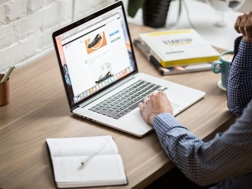 Eine Person arbeitet an einem Laptop an einem Schreibtisch mit Notizbuch, Tasse und Büchern. Der Laptop-Bildschirm zeigt ein Schuhdesign.