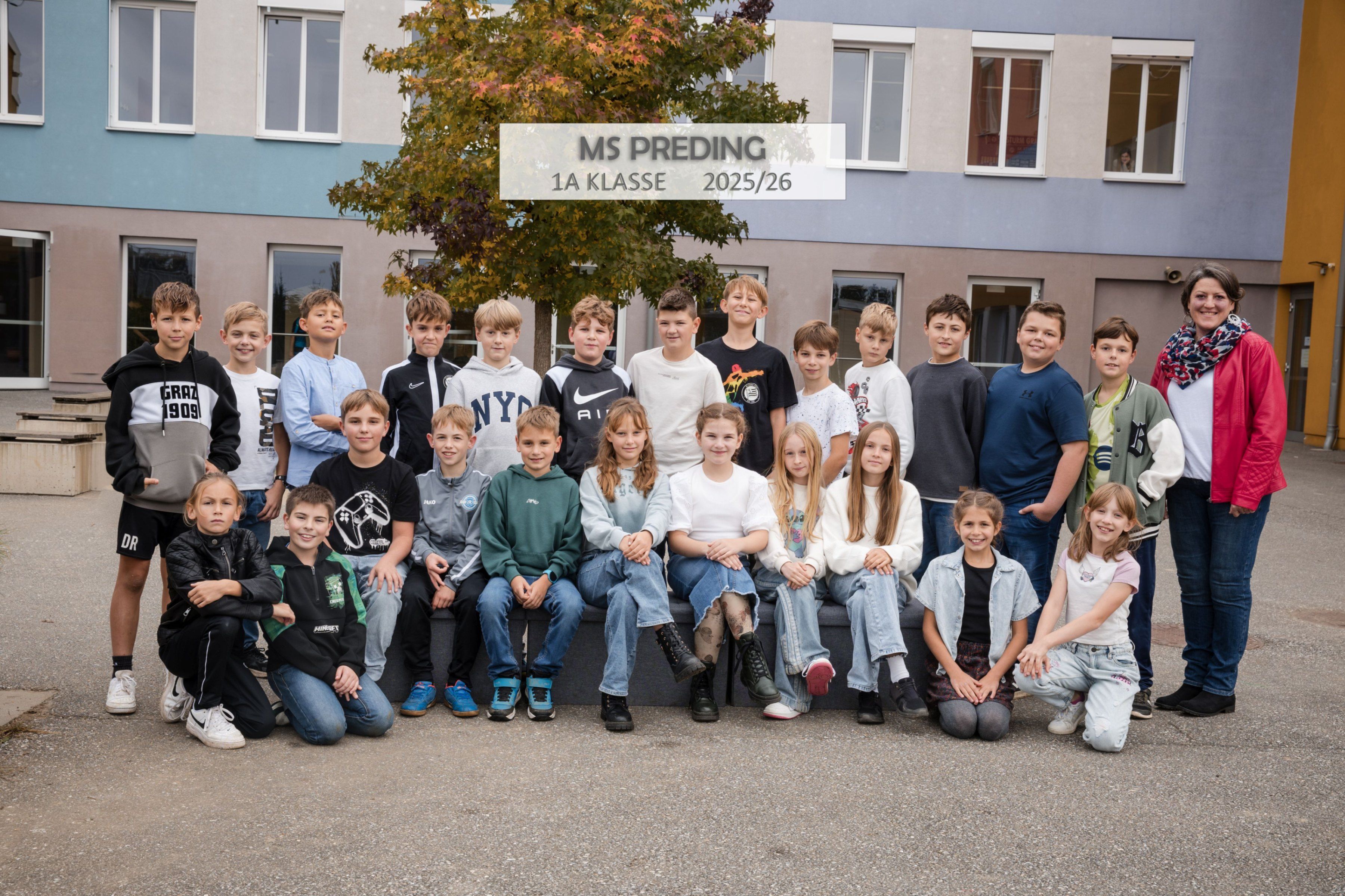 Gruppenfoto der Schüler der 1A Klasse von MS Preding im Jahr 2025/26. Sie sitzen auf einer Bank vor einem Gebäude mit einem Baum im Vordergrund.