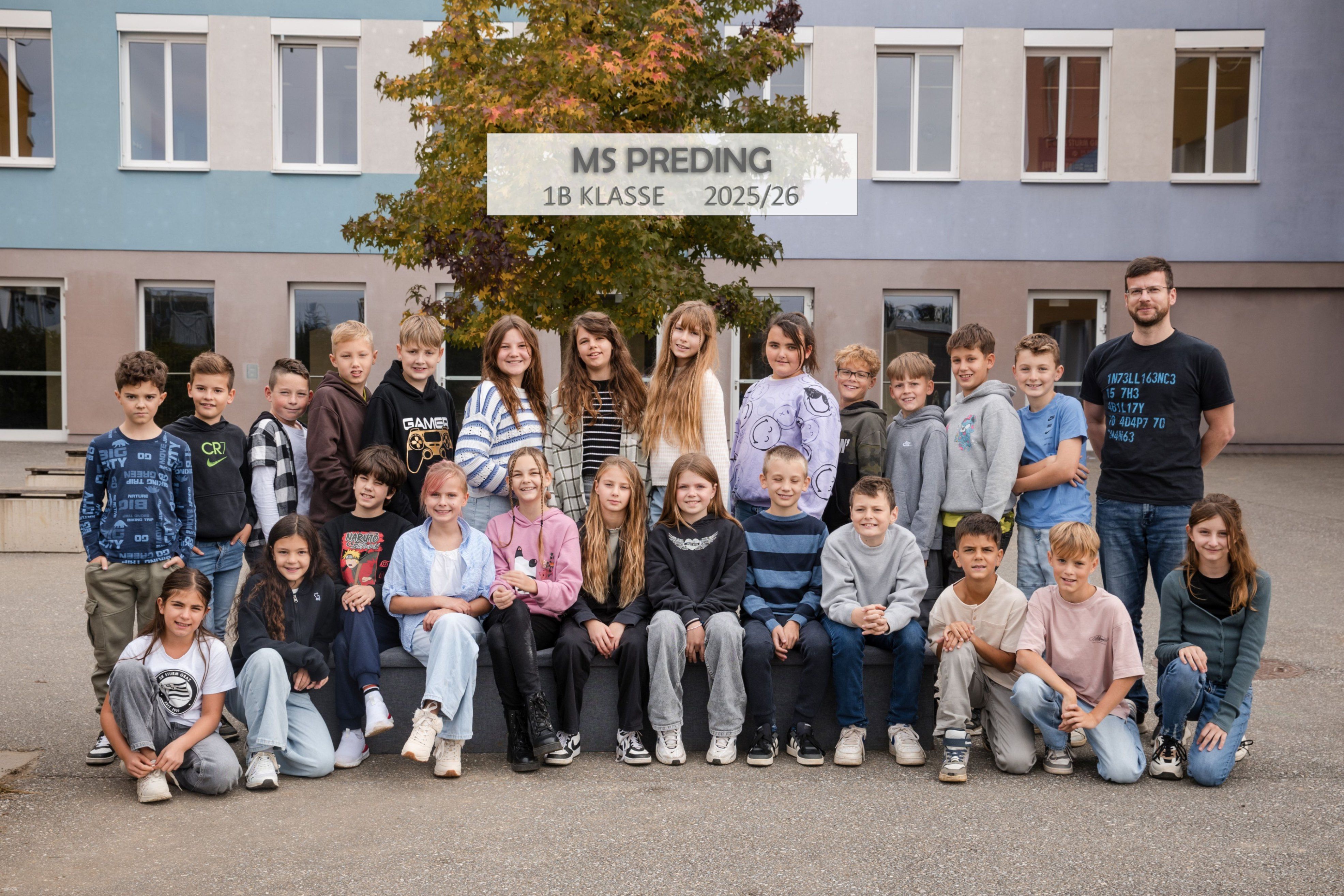 Eine Gruppe von Kindern posiert für ein Foto vor einem Schulgebäude. Darauf steht ein Schild MS Preding 1B Klasse 2025/26.