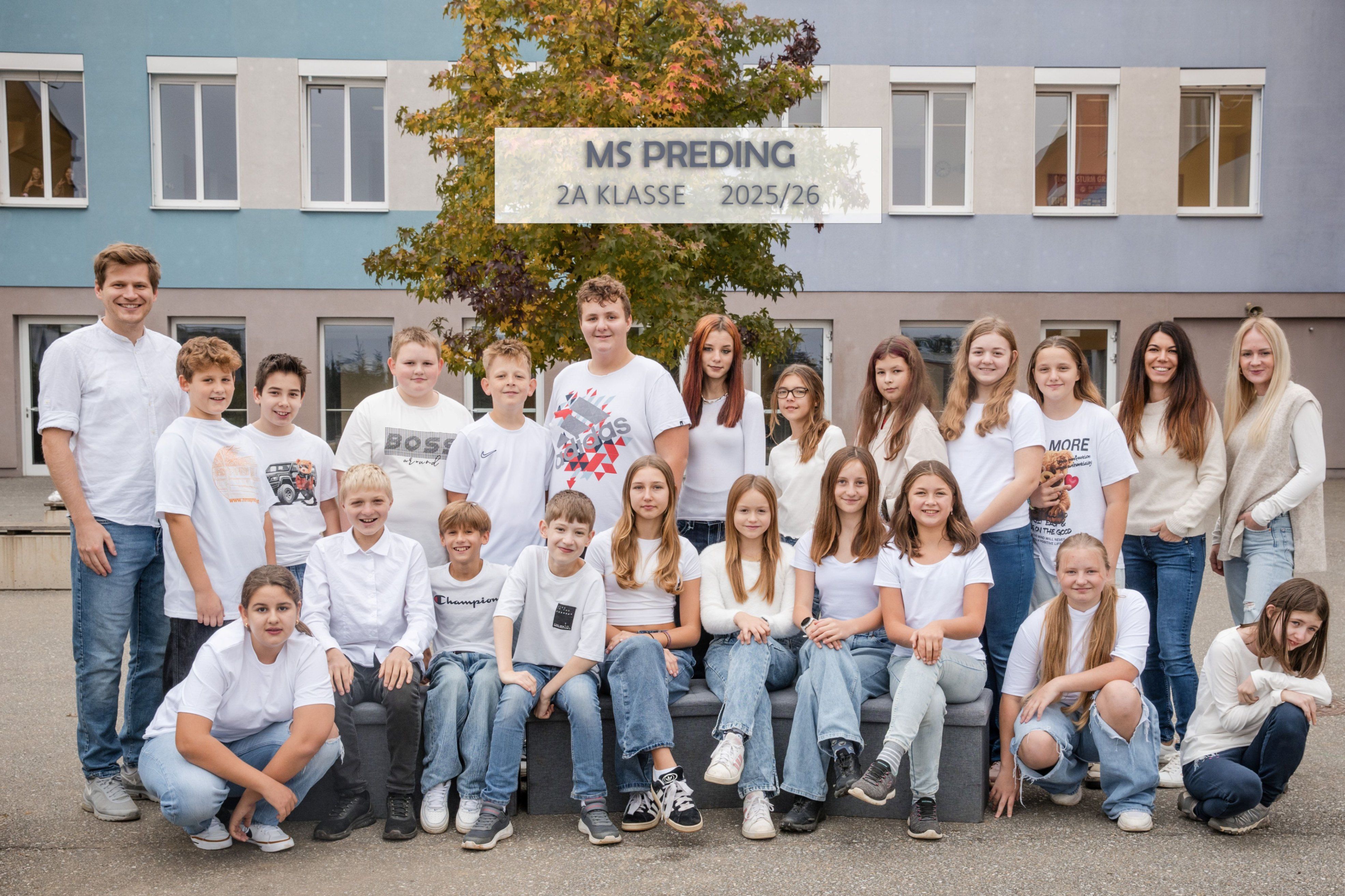 Eine Gruppe von Schülern der 2A Klasse posiert für ein Foto vor einem Gebäude, alle tragen weiße T-Shirts mit verschiedenen Aufdrucken.