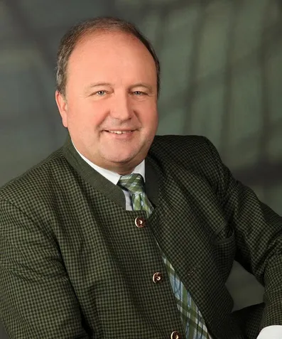 Ein Mann mit einem karierten Anzug und einer Krawatte lächelt in die Kamera. Er hat kurze Haare und trägt ein weißes Hemd. Er steht vor einem grünen Hintergrund.
