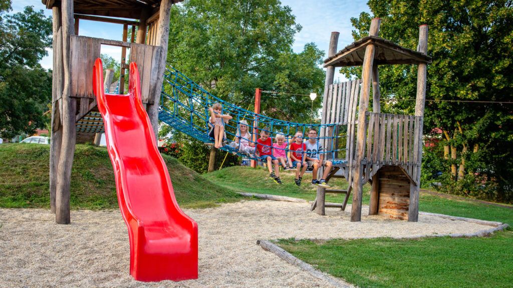 Kinder spielen auf einem Spielplatz mit einer roten Rutsche, einem Kletternetz und einer Holzkonstruktion.