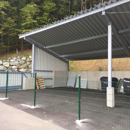 Ein leerer Carport mit Metalldach und grünem Zaun, umgeben von einer Steinmauer und Bäumen, mit zwei Mülltonnen in der Nähe.