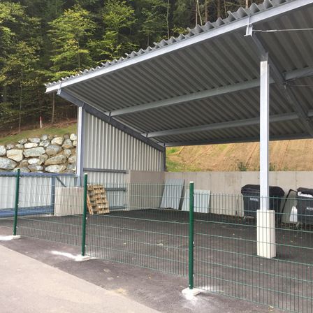 Ein leerer Carport mit Metalldach und grünem Zaun, umgeben von einer Steinmauer und Bäumen, mit zwei Mülltonnen in der Nähe.