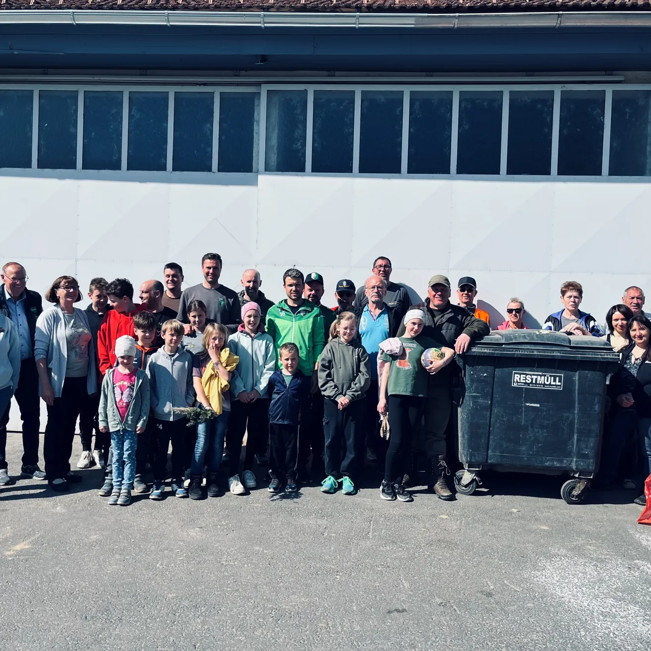 Eine Gruppe von Erwachsenen und Kindern steht vor einem Gebäude mit großen Fenstern. Ein schwarzer Mülleimer steht rechts. Sie scheinen für ein Gruppenfoto zu lächeln.