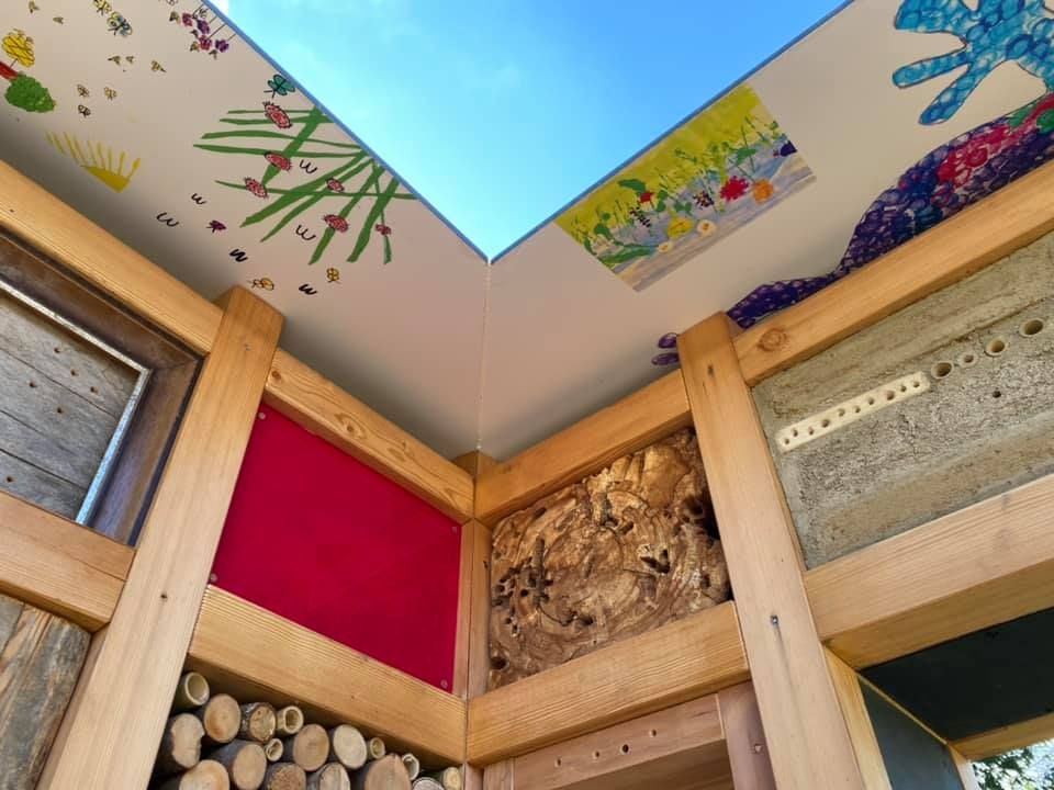 Nahaufnahme eines Innenraums aus Holz mit einer roten Wand, einem Fenster und einer offenen Holztür, die filigrane Schnitzereien, bemalte Wände und ein Oberlicht zeigt.