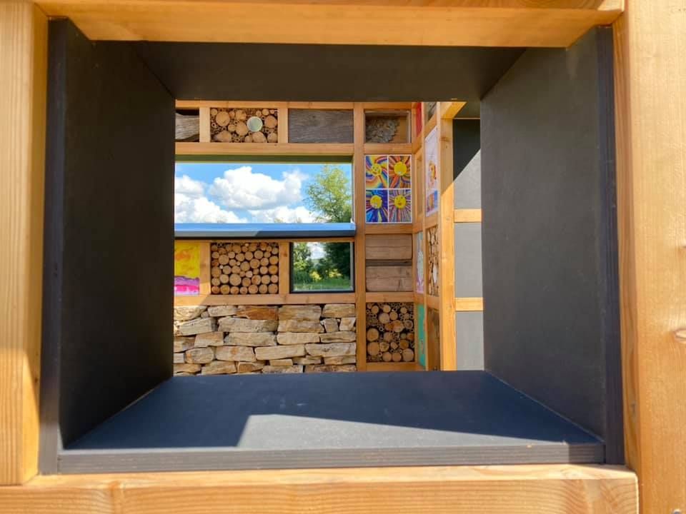 Innenansicht einer Holzstruktur mit schwarzem Rahmen und grauem Boden. Sie hat drei Fenster mit Holzrahmen, von denen eines eine Holzstange an der Seite hat. Es gibt auch eine Wand mit Gemälden und eine Steinmauer.