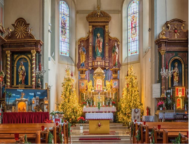 Ein Altar in einer Kirche, der für Weihnachten dekoriert ist. Auf beiden Seiten des Altars stehen zwei Weihnachtsbäume. Hinter dem Altar befinden sich zwei Statuen. Auf der linken Seite steht eine Krippe.