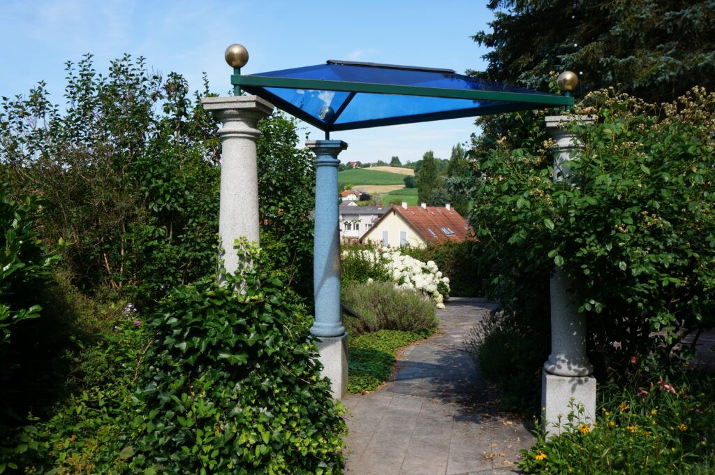 Ein blaues Pavillondach in einem Garten mit Pflanzen und einem Weg, der zu einem Haus mit rotem Dach führt.