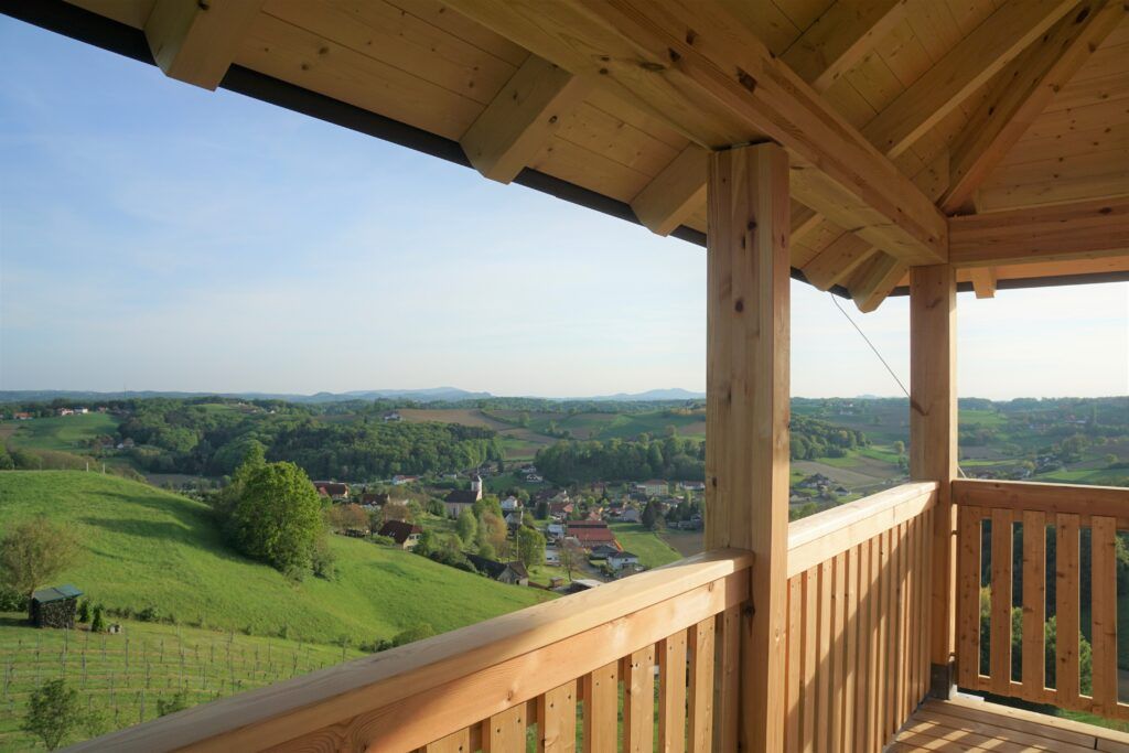 Ein Balkon mit einem hölzernen Geländer bietet einen Panoramablick auf ein grünes Tal, Hügel und ein Dorf unter einem klaren blauen Himmel.