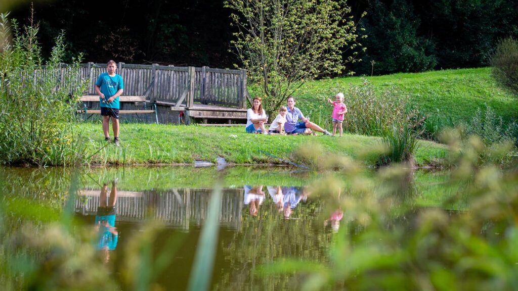 Eine Familie genießt einen Tag im Freien, wobei eine Person im Teich fischt, während andere auf dem Gras sitzen.