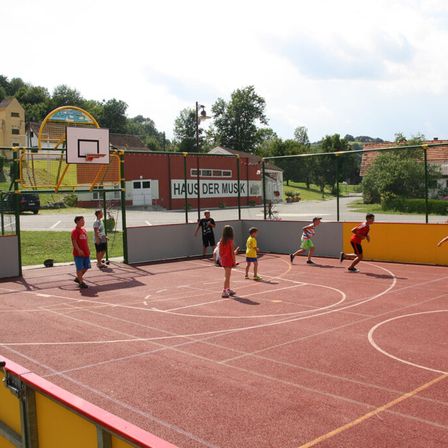 Mehrere Kinder spielen Basketball auf einem Freiluftplatz. Ein Basketballkorb befindet sich am Ende des Platzes.