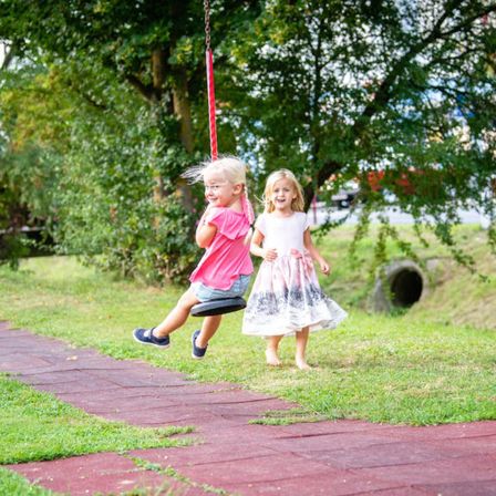 Drei junge Mädchen sind in einem Park. Eine steht auf dem Boden, eine andere schwingt auf einer Schaukel und die dritte geht auf die Schaukel zu.