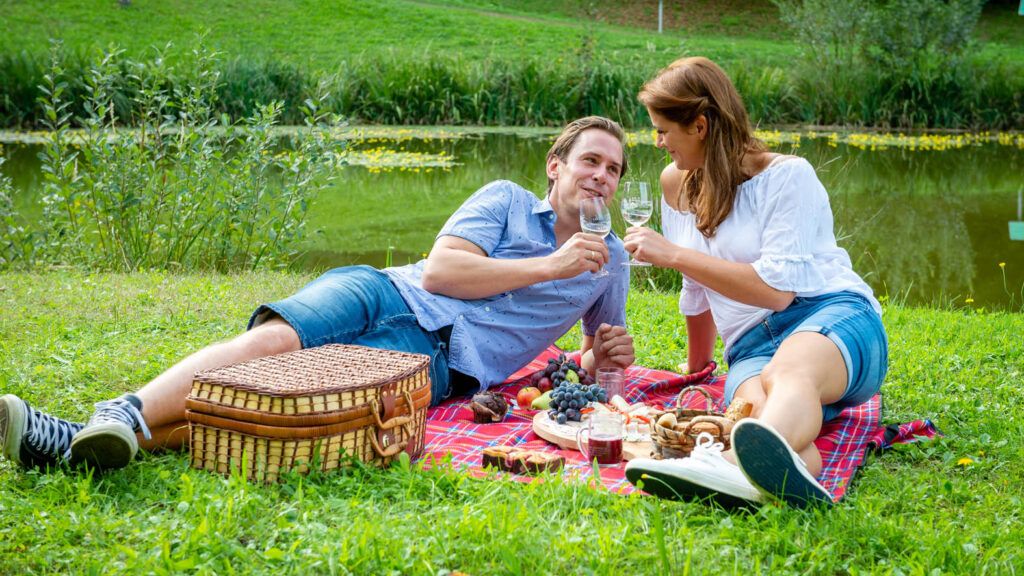 Ein Mann und eine Frau machen ein Picknick auf der Wiese am See. Sie lächeln und trinken Wein.