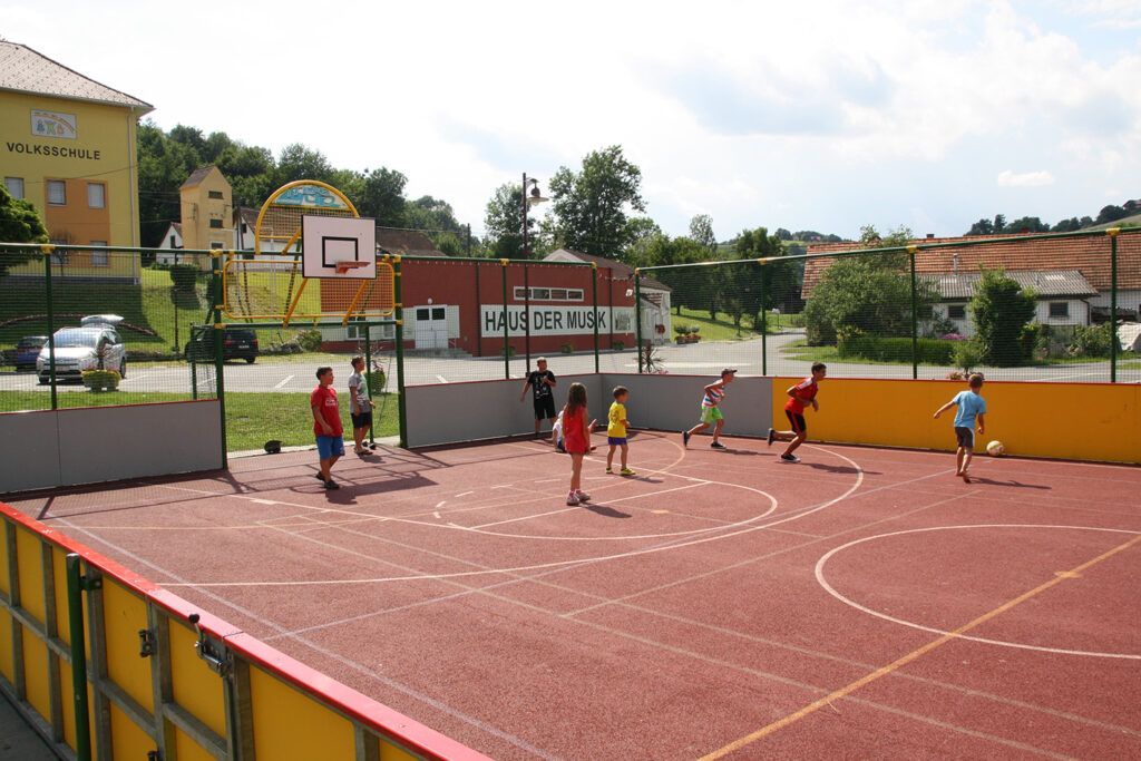 Mehrere Kinder spielen Basketball auf einem Freiluftplatz. Ein Basketballkorb befindet sich am Ende des Platzes.