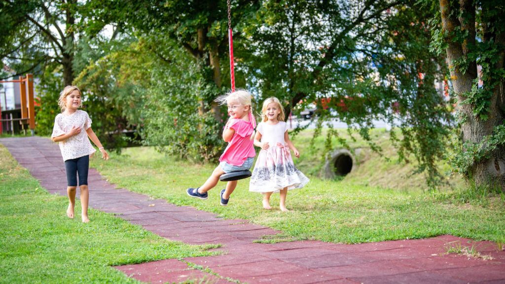 Drei junge Mädchen sind in einem Park. Eine steht auf dem Boden, eine andere schwingt auf einer Schaukel und die dritte geht auf die Schaukel zu.