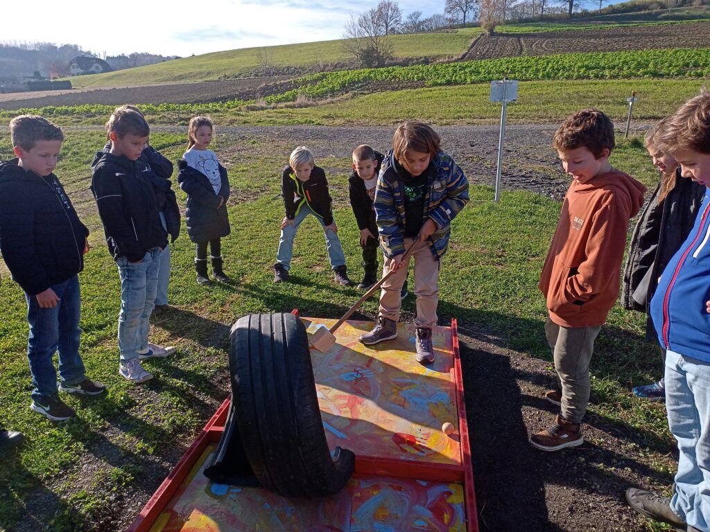 Gruppe von Kindern spielt auf einer Reifenschaukel auf einem Feld mit einer Kulisse eines Hügels. Ein Kind benutzt einen Hammer, um einen Ball zu schlagen.