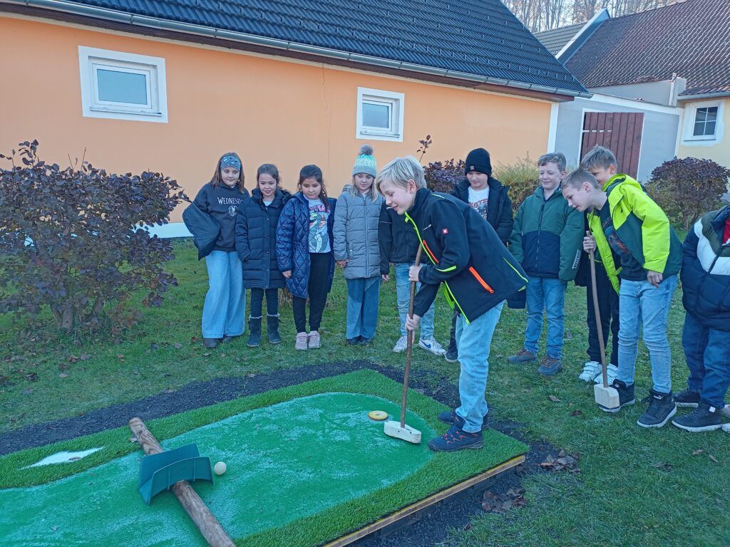 Eine Gruppe von Kindern spielt Minigolf im Garten eines Hauses. Ein Kind schlägt den Ball.