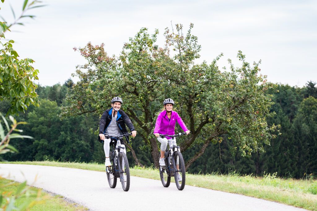 Zwei Personen fahren auf einem Weg umringt von Bäumen und Grünflächen mit Fahrrädern.