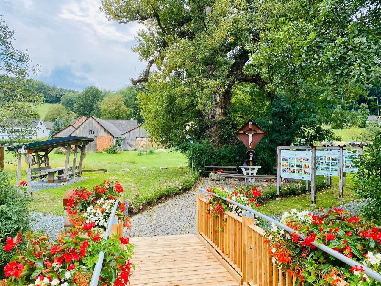 Ein Blick auf einen Holzweg mit einer Bank auf der linken Seite und Blumen auf der rechten Seite. Ein Holzhaus und ein Kruzifix sind in der Ferne.