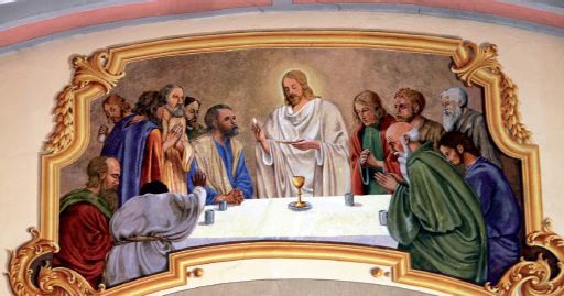 Eine Wandmalerei, die Jesus Christus und seine Jünger beim Letzten Abendmahl darstellt. Die zentrale Figur von Jesus ist prominent dargestellt, umgeben von seinen Aposteln, mit einem goldenen Kelch auf dem Tisch.