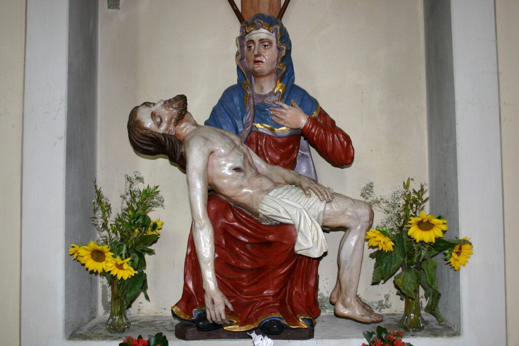 Eine Statue einer Frau, die einen toten Mann hält, steht in einer Nische. Die Frau trägt ein blaues und rotes Gewand, während der Mann ein weißes Tuch trägt. Auf beiden Seiten der Statue stehen Vasen mit Sonnenblumen.