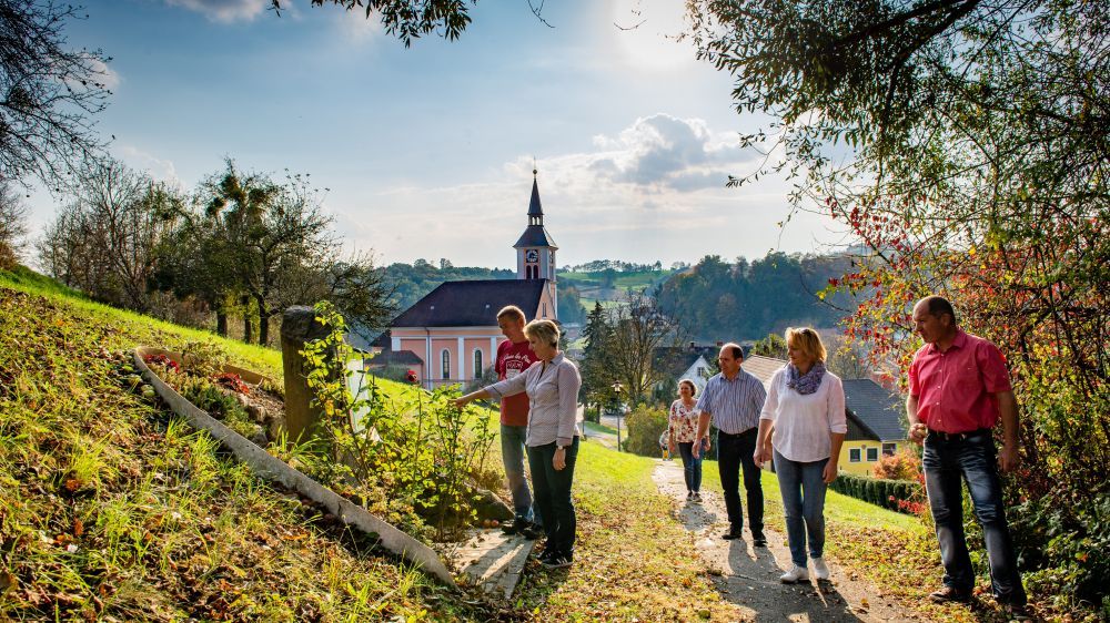 Eine Gruppe von Menschen geht auf einem Pfad. Sie laufen auf einem Weg mit einem Grasbereich auf der Seite. Sie gehen in Richtung einer Kirche mit Turm und Dachreiter. Dahinter befinden sich einige Häuser mit Zaun und Bäumen.
