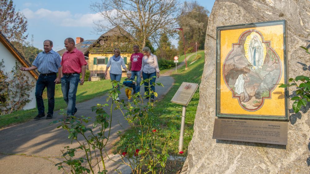 Eine fünfköpfige Familie läuft auf einem Pfad neben einem Haus und einem Schild, mit einer Felswand, die ein religiöses Gemälde im Hintergrund zeigt.