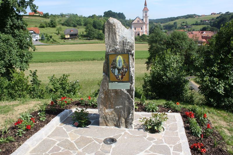 Ein großer Steinmonument mit einer Tafel befindet sich in einem Garten, umgeben von Bäumen und Blumen, mit einer Kirche im Hintergrund.