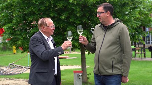 Zwei Männer, einer im Anzug und der andere in einer grünen Jacke, prosten mit Weinläsern in einer Außenumgebung an.