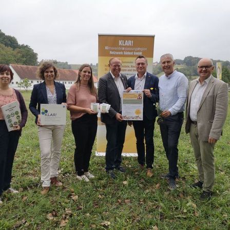 Sechs Menschen stehen auf einem Feld und halten Bücher und Broschüren in der Hand, sie lächeln für die Kamera. Hinter ihnen steht ein Banner mit der Aufschrift 'KLAR! Netzwerk Südsteiermark GmbH'. Im Hintergrund sind Häuser und Bäume zu sehen.